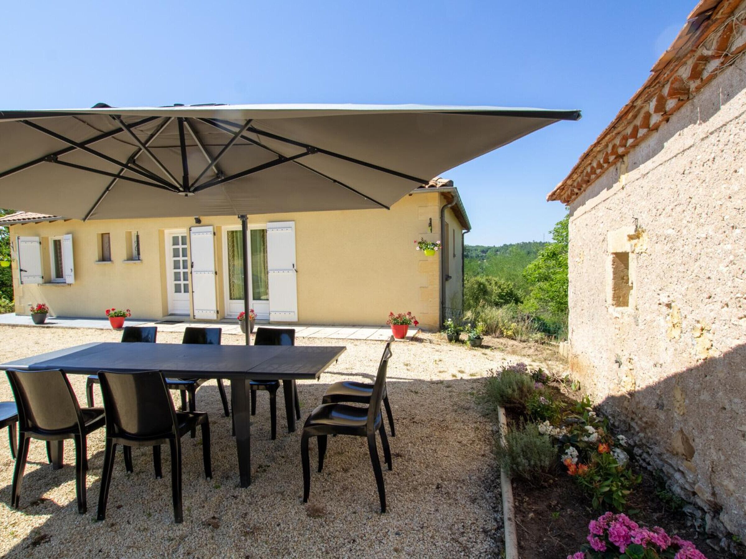 An outdoor patio with a table and chairs under a large umbrella, a building with a yellow facade, and a stone wall.