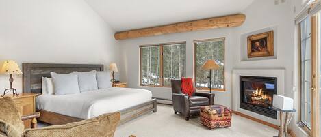 A bedroom with a large bed, a fireplace, a window with a view of trees, and a cozy chair.