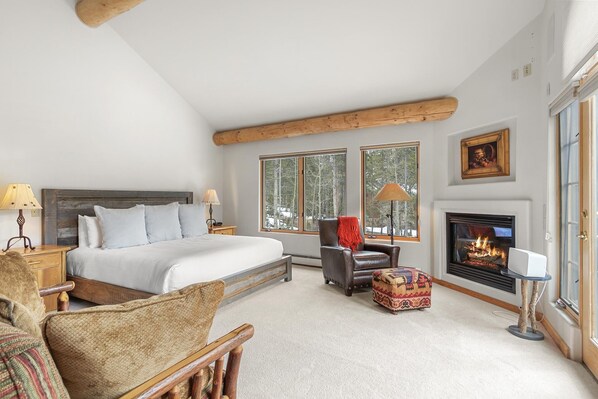 A bedroom with a large bed, a fireplace, a window with a view of trees, and a cozy chair.