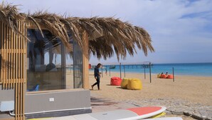 Un bâtiment situé au bord de la plage, doté d’un toit de chaume, d’une structure en verre et d’une planche de surf posée sur le sol.
