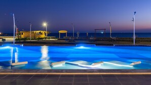 Une piscine au crépuscule, offrant une vue sur le coucher de soleil, entourée de chaises longues et d’une structure recouverte de chaume.