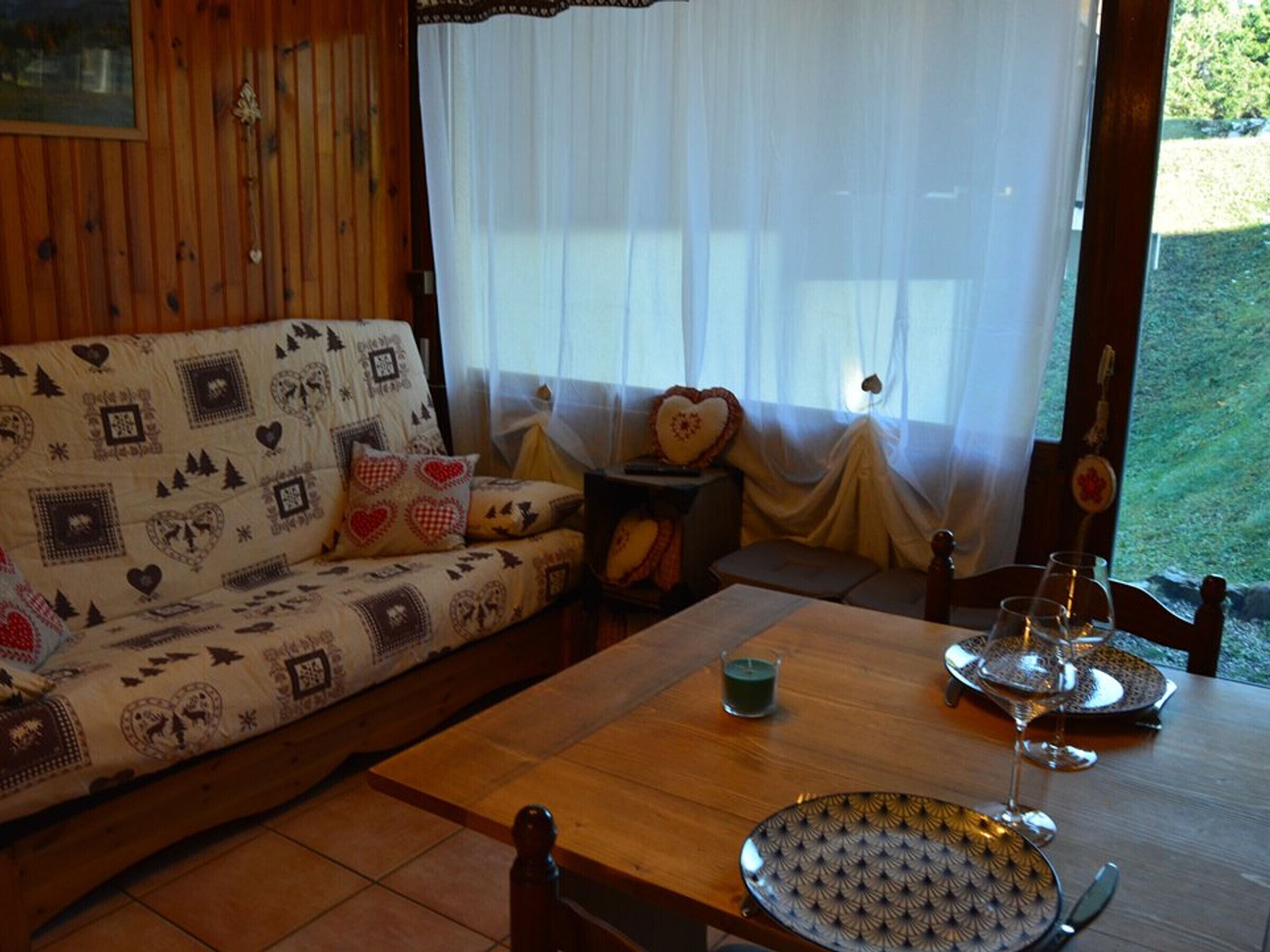 A cozy room with a wooden sofa adorned with heart patterns, a small table with a candle, and a view of greenery outside.