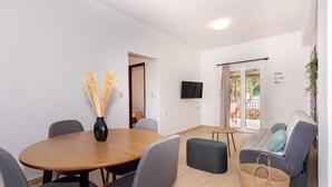 A dining area with a round wooden table, grey upholstered chairs, a black vase with dried plants, a small round table with a chair, a grey sofa with cushions, and a wicker basket. - Mysa Nature Apartments (Zakynthos)