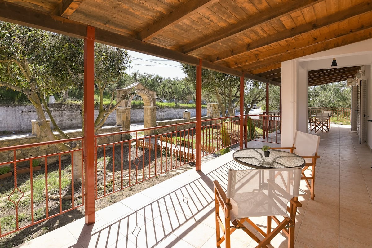 Eine überdachte Terrasse im Freien mit Tisch und Stühlen, einer Steinwand und Bäumen im Hintergrund.