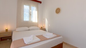 A neatly made bed with white linens and a beige quilt, two bedside tables with lamps, a large window with sheer curtains, and a circular wall decoration. - Mysa Nature Apartments (Zakynthos)