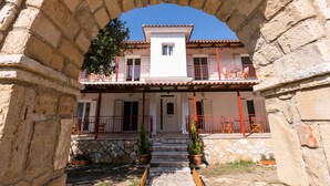 A two-story building with a stone archway entrance, a balcony with red railings, and a small garden area with potted plants. - Mysa Nature Apartments (Zakynthos)