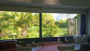 Une pièce avec une table et des chaises, une fenêtre donnant sur un paysage verdoyant, et divers objets placés sur le rebord de la fenêtre.