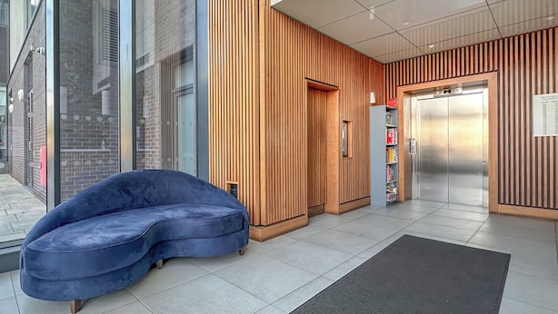 Un hall d’entrée moderne avec un canapé bleu incurvé, des panneaux de bois, un ascenseur et une bibliothèque.