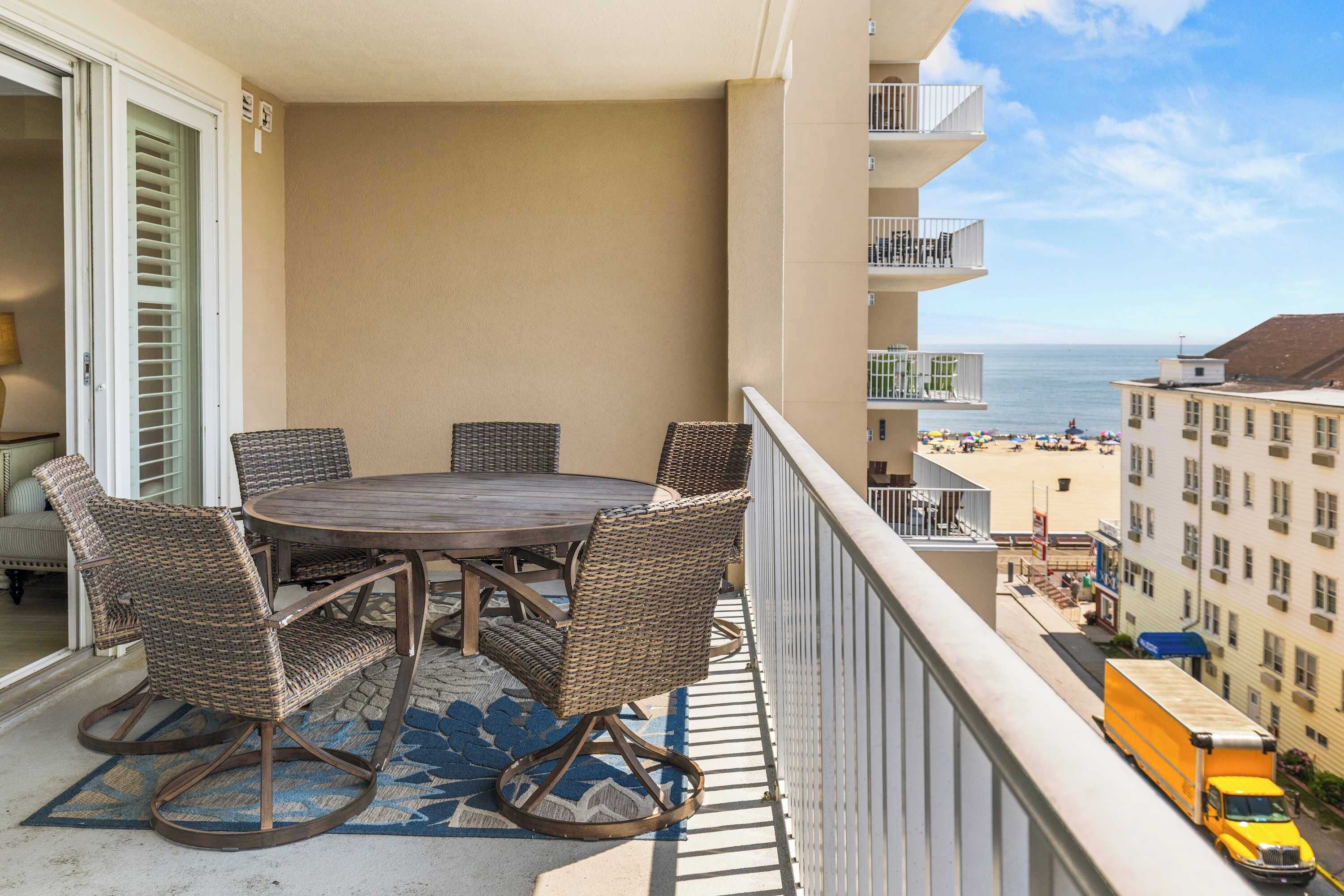 A balcony with wicker furniture, a view of the beach, and a building across the street.