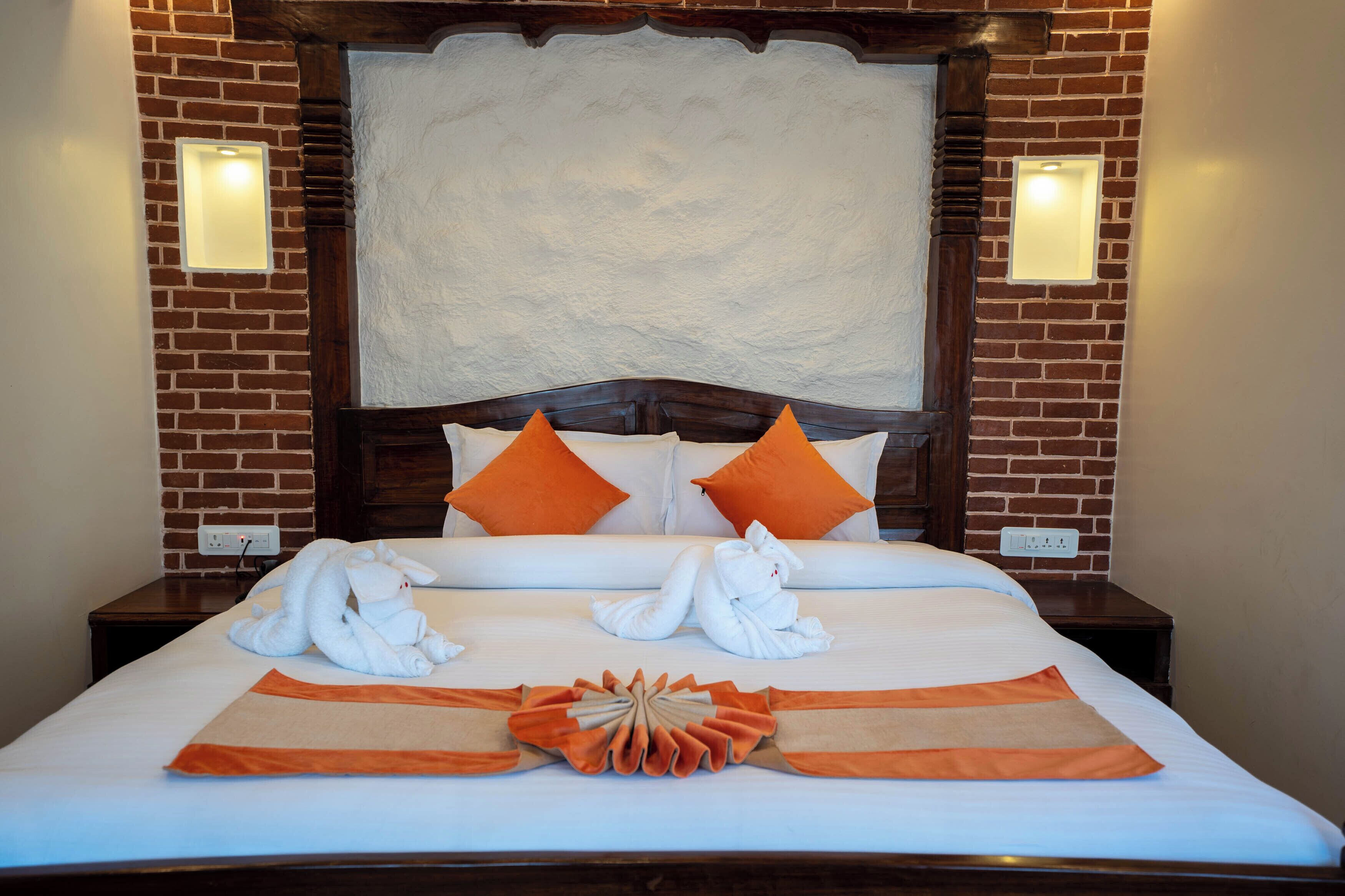 A hotel room with a brick wall, a large bed with white bedding, orange pillows, and two white towels shaped like animals. There are two wall-mounted lights and a power outlet on the wall.