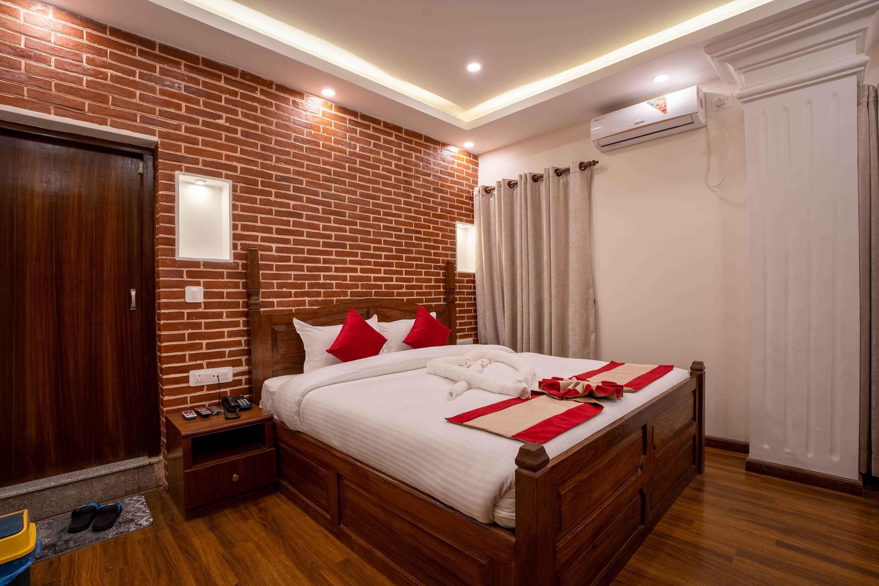 Una habitación de hotel con pared de ladrillos, cama de madera con almohadas rojas, unidad de aire acondicionado blanca y puerta de madera.