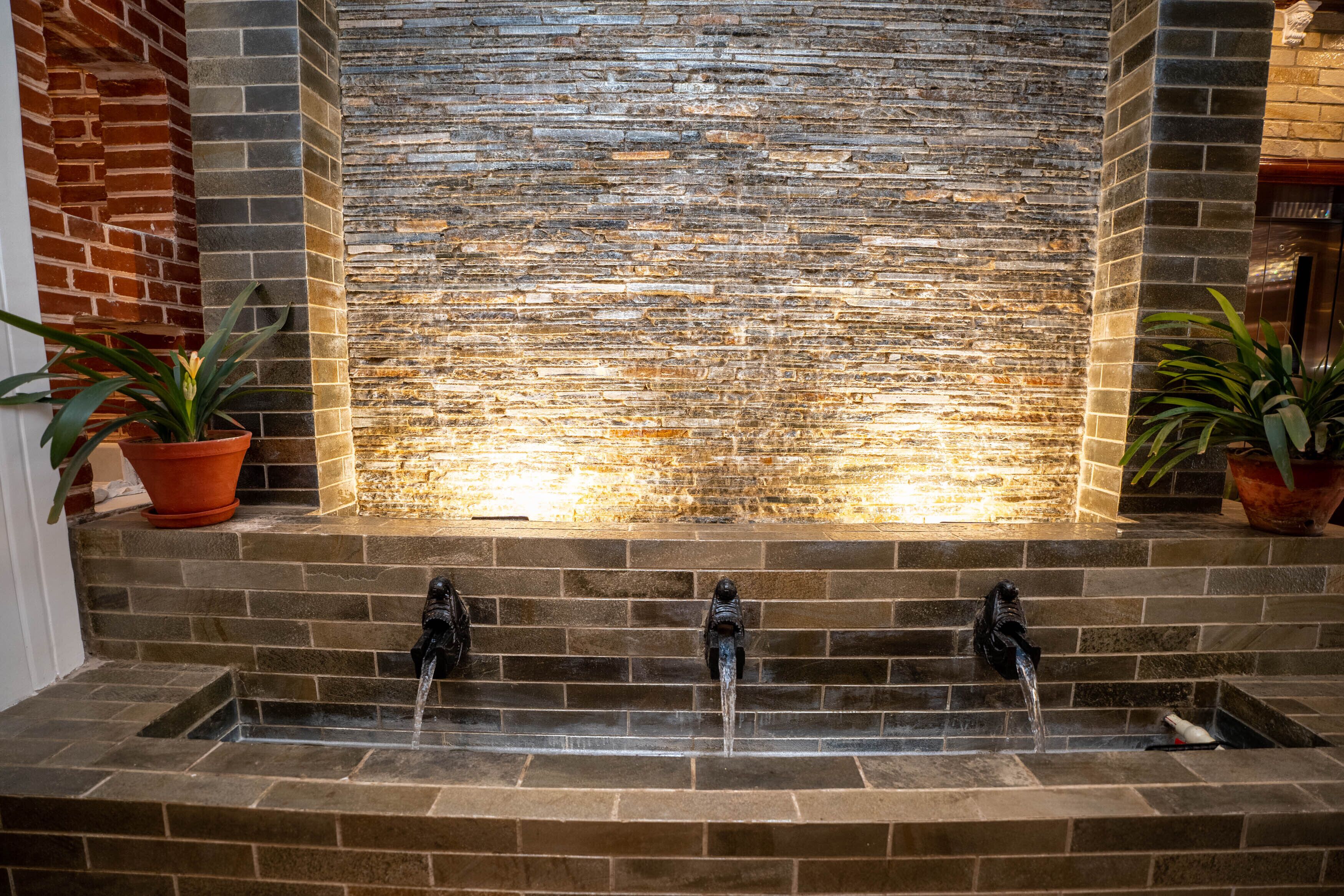 A stone water feature with three spouts, flanked by brick walls and potted plants.