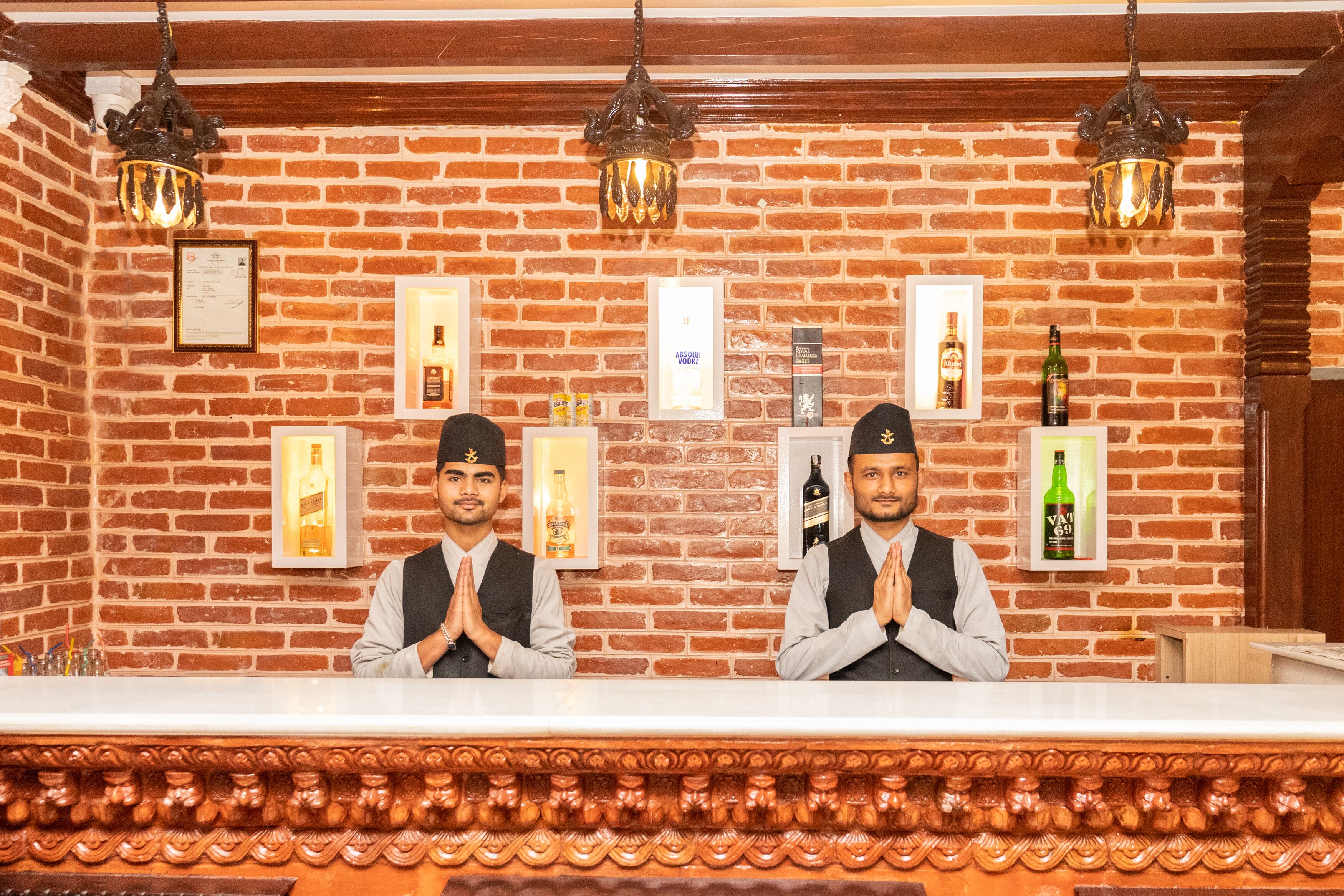 Un área de recepción con dos empleados, una pared de ladrillo, cuadros enmarcados y varias botellas de alcohol.