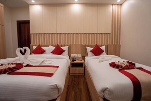 Habitación de hotel con dos camas, cada una con ropa blanca y almohadas rojas, un cabecero de madera, una mesita de noche pequeña con una lámpara, y suelo de madera.