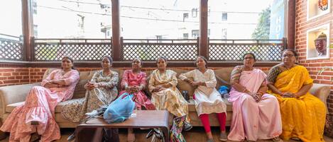Un grupo de mujeres sentadas en un sofá en una habitación con suelo de madera y paredes de ladrillo, una ventana con vista a edificios y una decoración colgante.