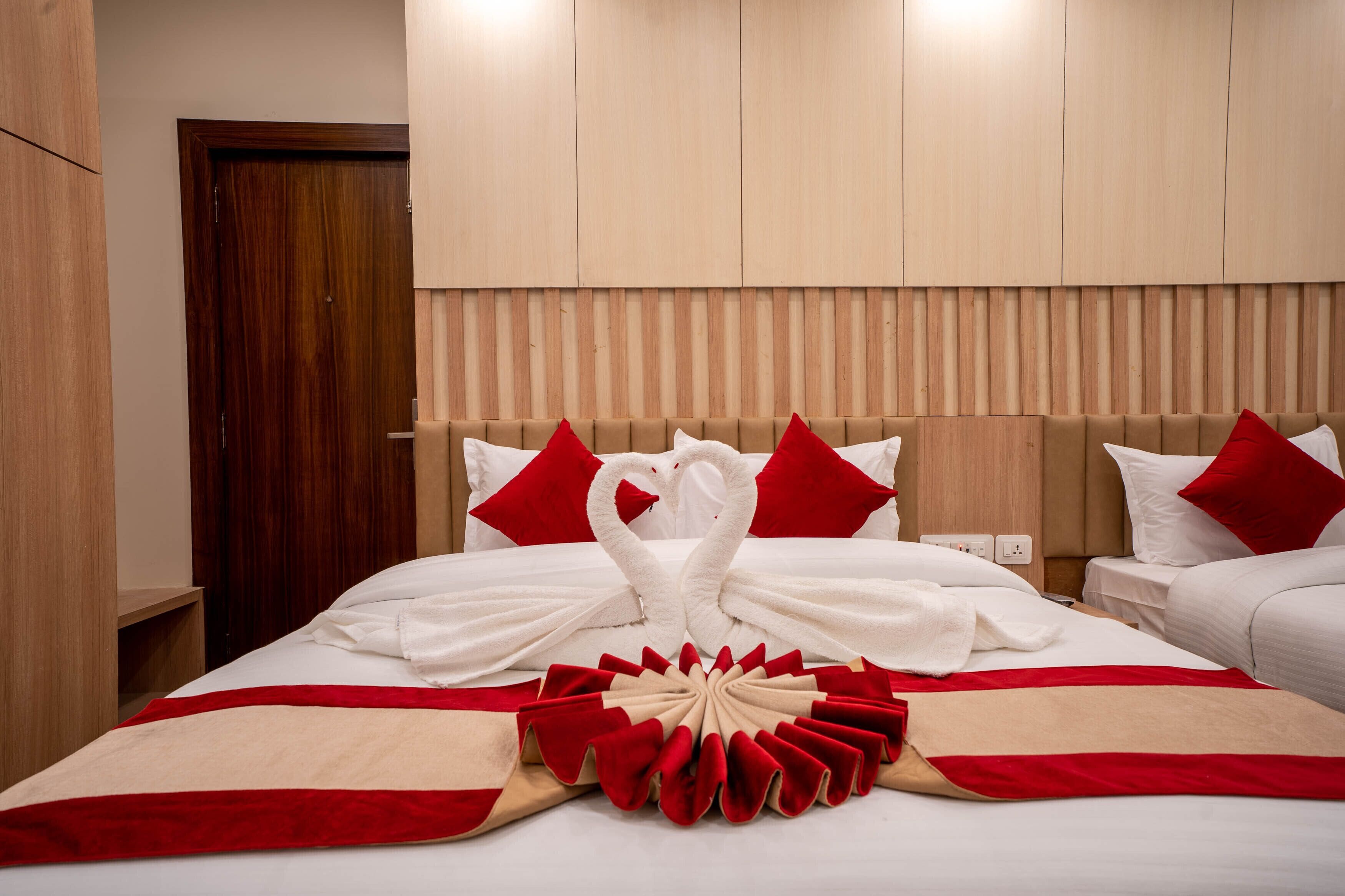 A hotel room with a bed featuring a red and white swan towel arrangement, a red and beige decorative pillow, and a wooden headboard.