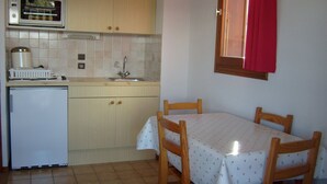 Eine kleine Küche mit Mikrowelle, Spüle und kleinem Kühlschrank. Ein Esstisch mit Stühlen und ein Fenster mit roten Vorhängen sind vorhanden.