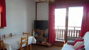 Ein Zimmer mit Esstisch, Stühlen, Fernseher, Sofa und einem Balkon mit roten Vorhängen.