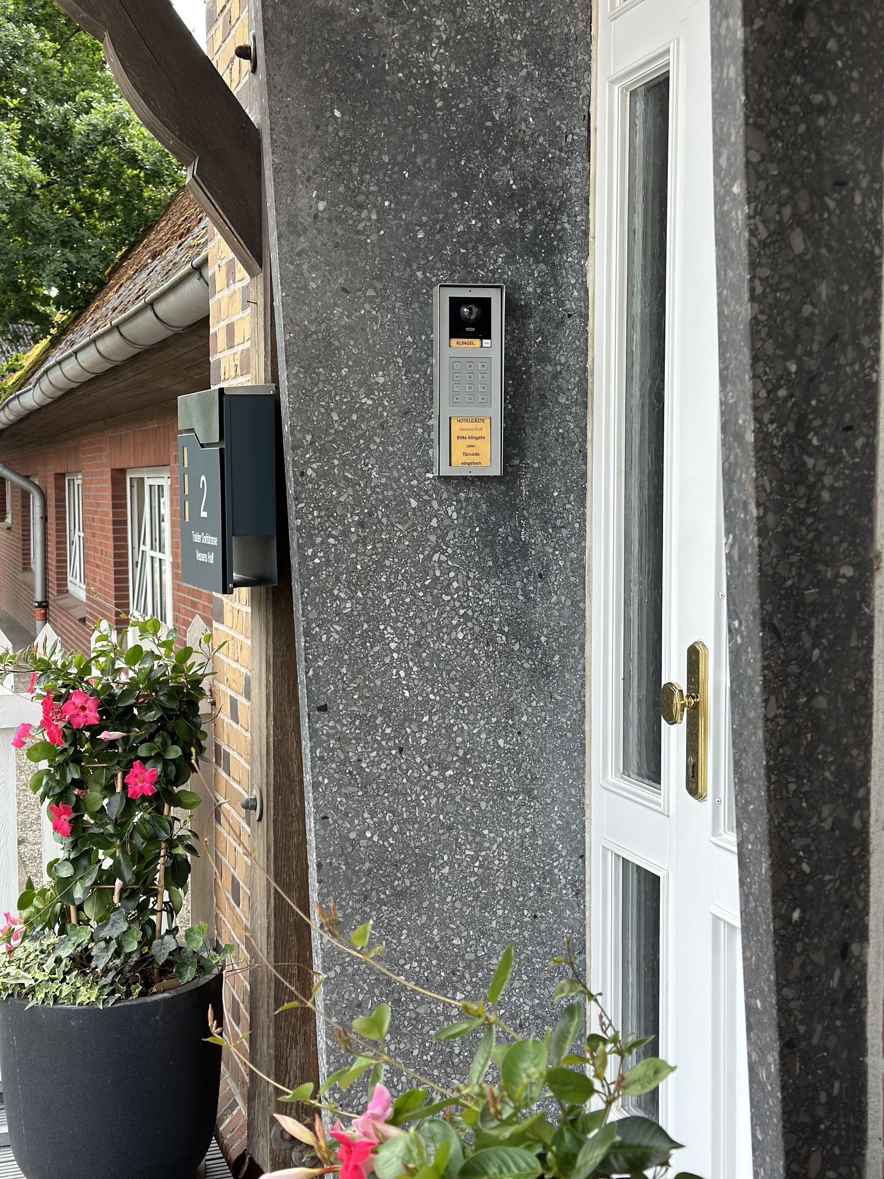 Un bâtiment doté d’une porte, d’un interphone fixé au mur et d’une plante en pot ornée de fleurs.