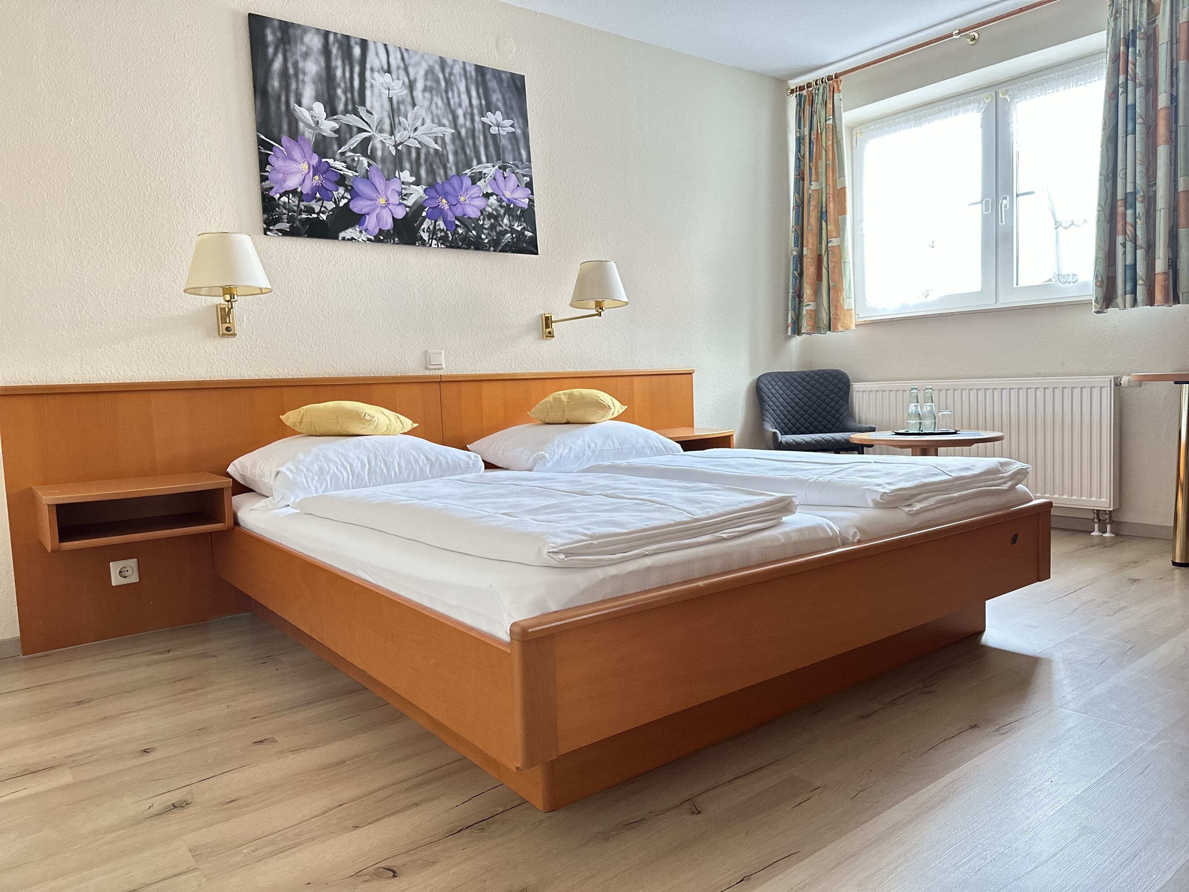 A hotel room with a wooden bed, two bedside lamps, a nightstand, a chair, a table, and a window with curtains.