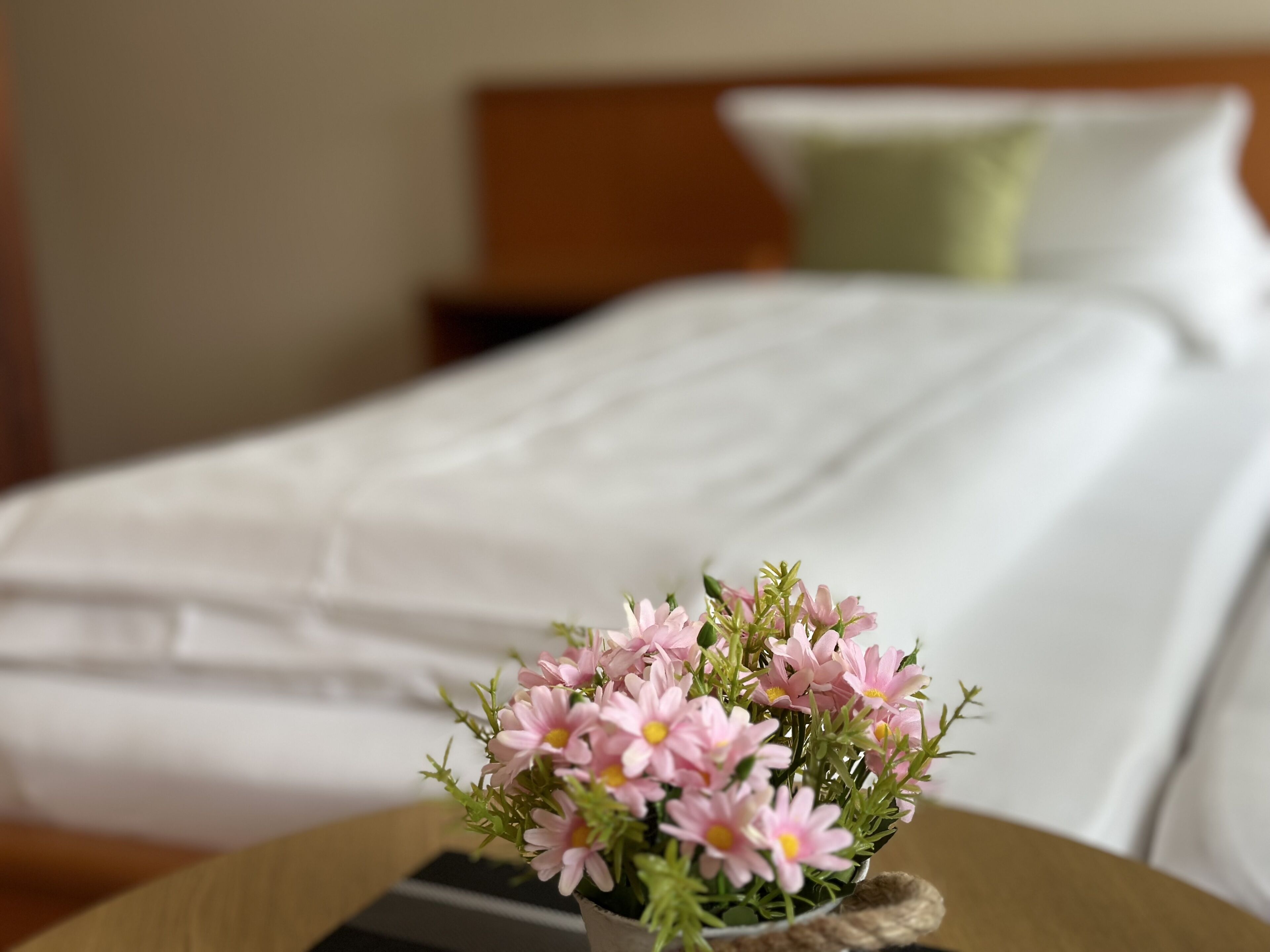 Una cama bien hecha con sábanas blancas y una almohada verde al fondo. En primer plano, una pequeña mesa de madera con un jarrón de flores rosas.