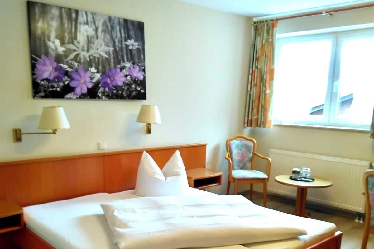 A hotel room with a large bed, two bedside lamps, a small table, a chair, and a window with curtains.