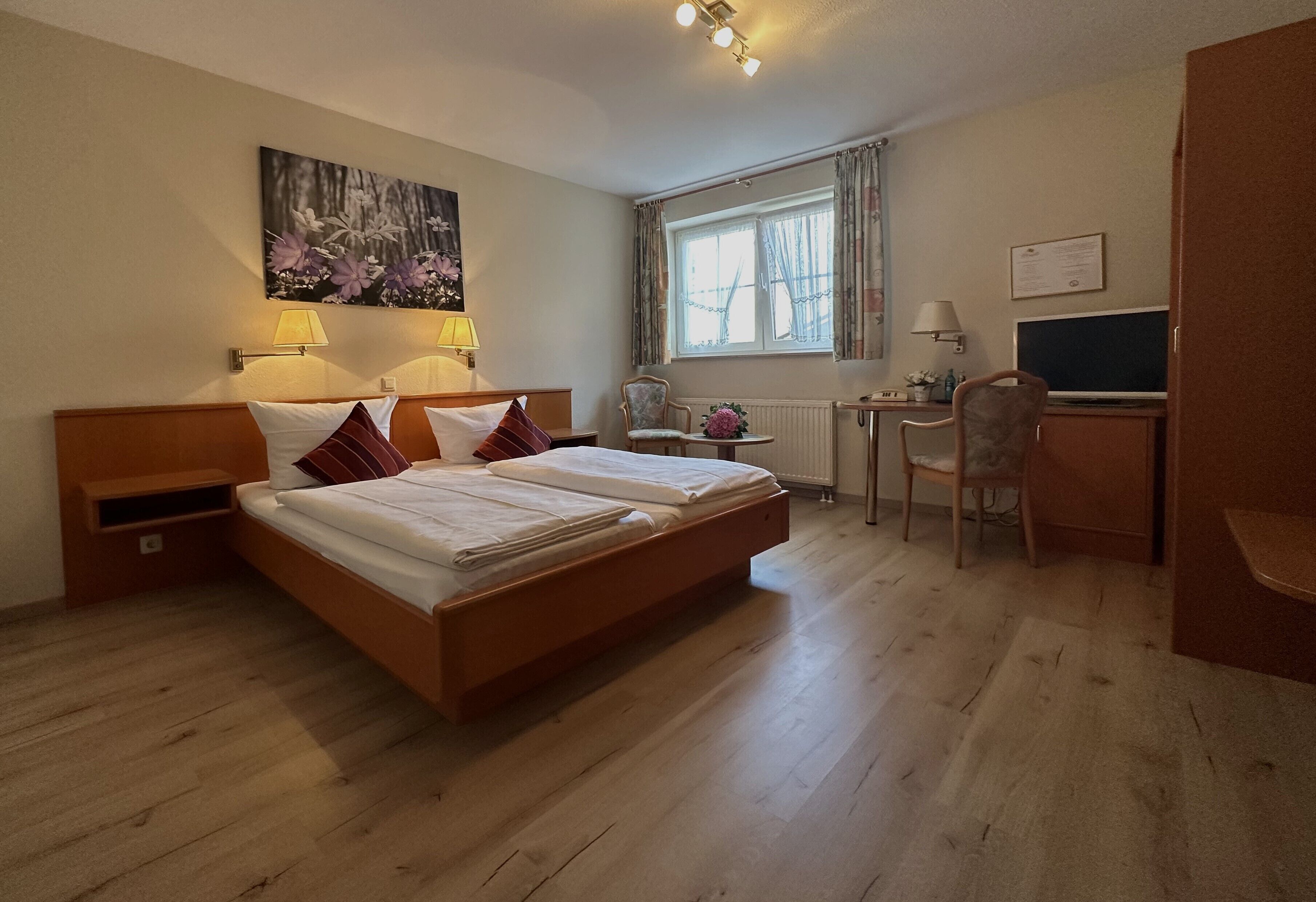A hotel room with a double bed, two bedside lamps, a desk with a chair, a window with curtains, and a wall-mounted TV.