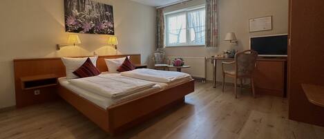 A hotel room with a double bed, two bedside lamps, a desk with a chair, a window with curtains, and a wall-mounted TV.
