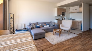 A modern living room with a grey sofa, a white armchair, and a small wooden side table. A kitchen area is visible in the background with wooden cabinets and a microwave. - "Charming Studio in Central Arcachon with Balcony, Steps from Beach - Sleeps 2" (Arcachon)