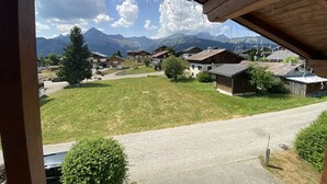 En vy från en träbalkong med utsikt över en by med traditionella hus, en gräsyta och berg i bakgrunden.