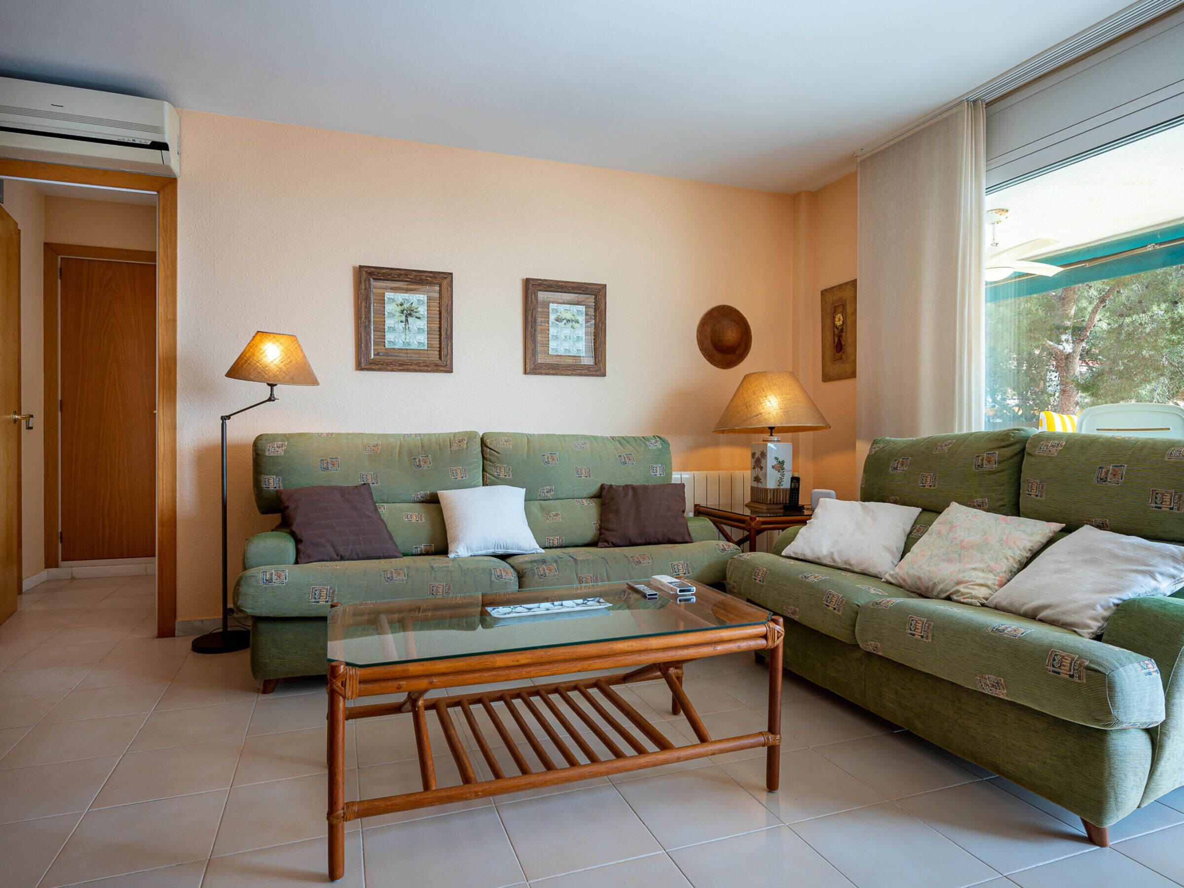 A living room with a green sofa set, a glass-top coffee table, two wall-mounted lamps, and a view through a window.