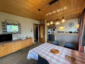 Um quarto aconchegante com teto de madeira, televisão, mesa de jantar com toalha xadrez e cozinha equipada com relógio e micro-ondas.