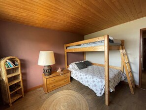 Uma cama beliche com escada de madeira, uma mesa de cabeceira com abajur, e uma prateleira de vime com livros.