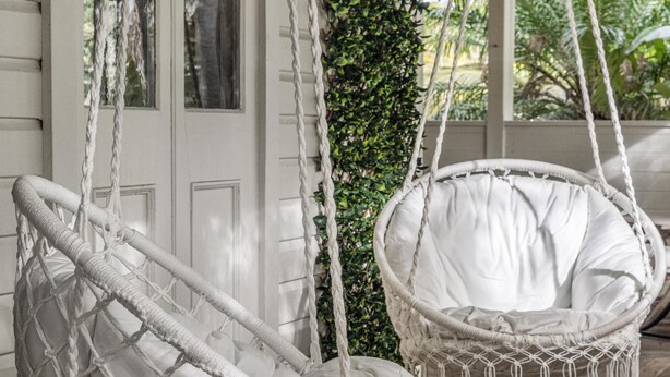 Une chaise suspendue en osier blanc, dotée d’un coussin, placée devant une porte blanche et un mur recouvert de verdure.