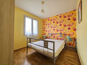 Un dormitorio con estructura de cama metálica, dos mesitas de noche con lámparas, una ventana con cortina translúcida y papel tapiz con estampado floral.