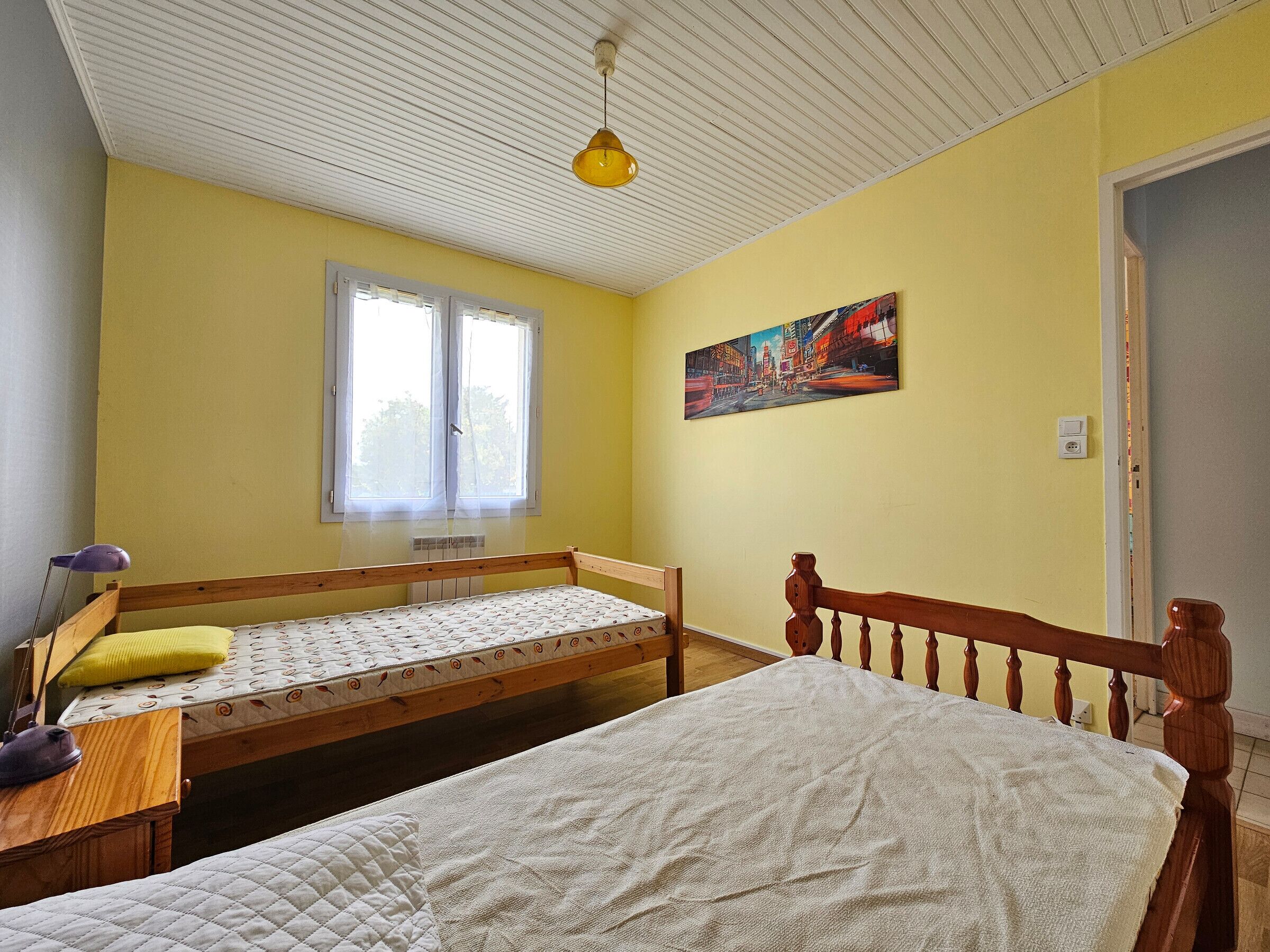 A room with two wooden beds, a window with white curtains, a yellow wall, and a painting.
