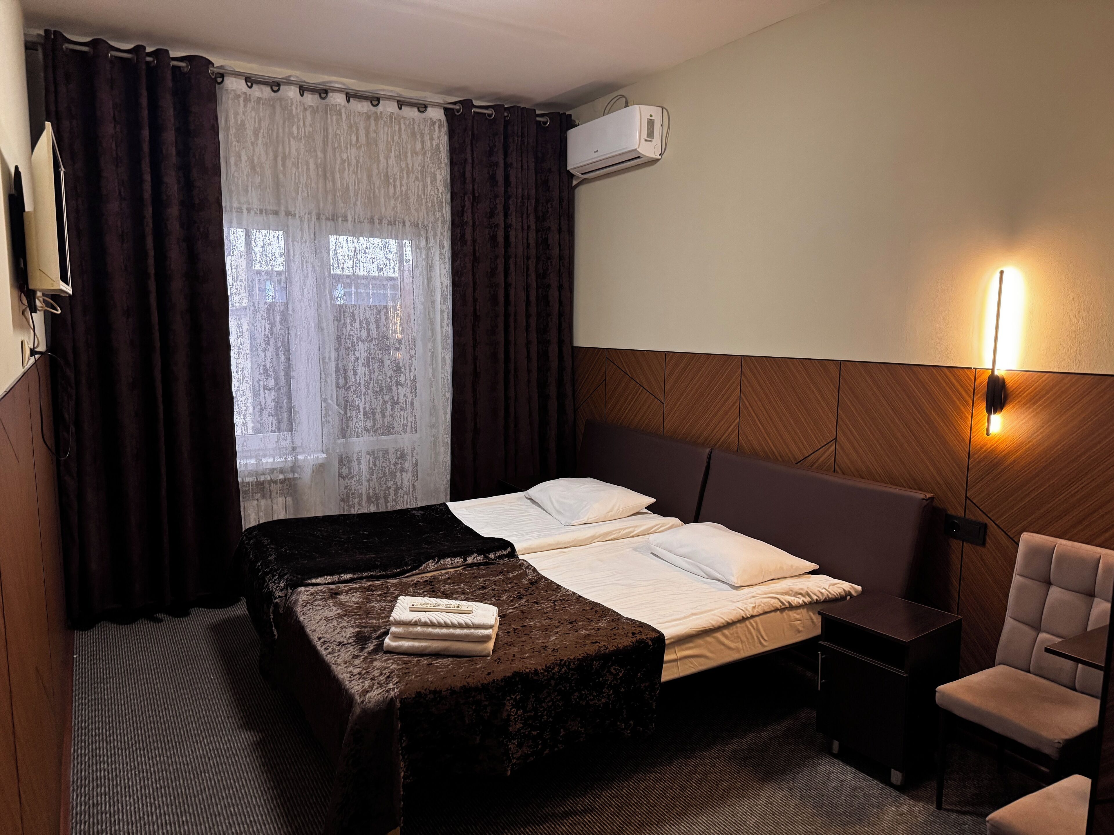 Une chambre d’hôtel avec un lit, une petite table, une chaise, un appareil de climatisation et une fenêtre avec des rideaux.