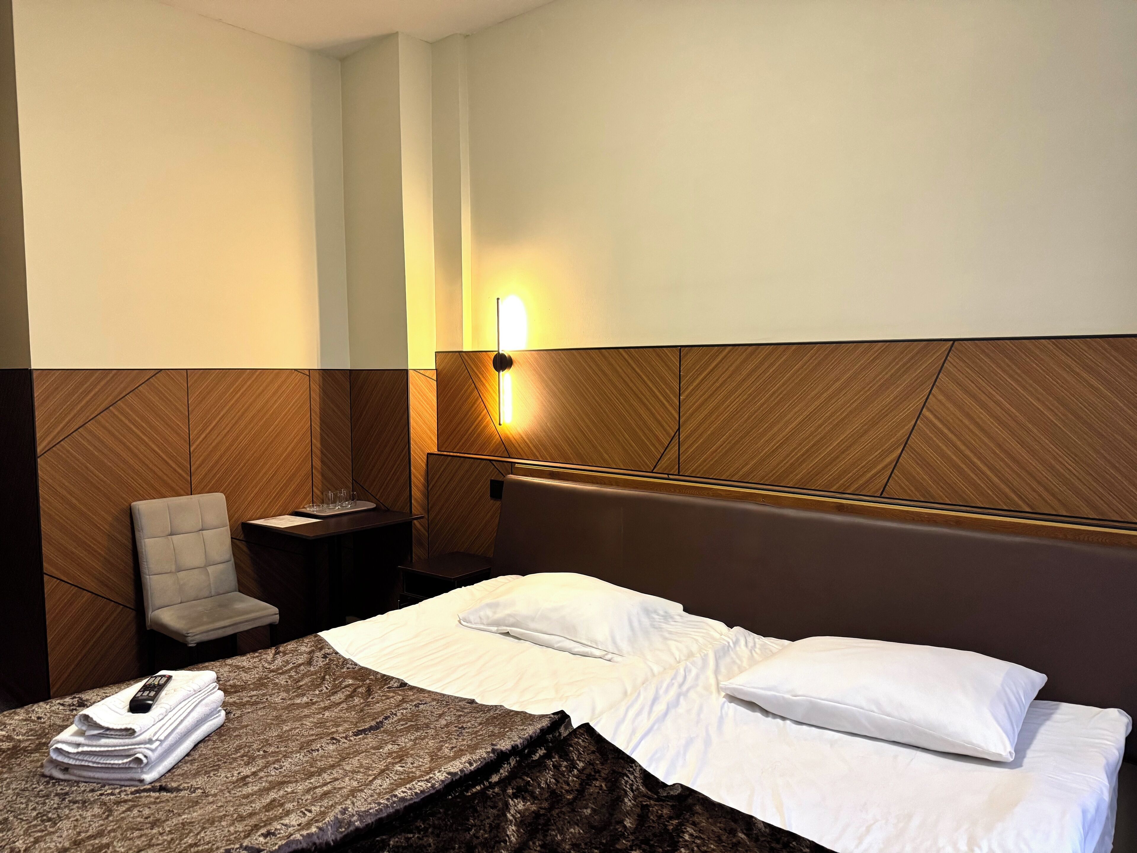 A hotel room with a bed, a chair, a nightstand, and a wall-mounted light.