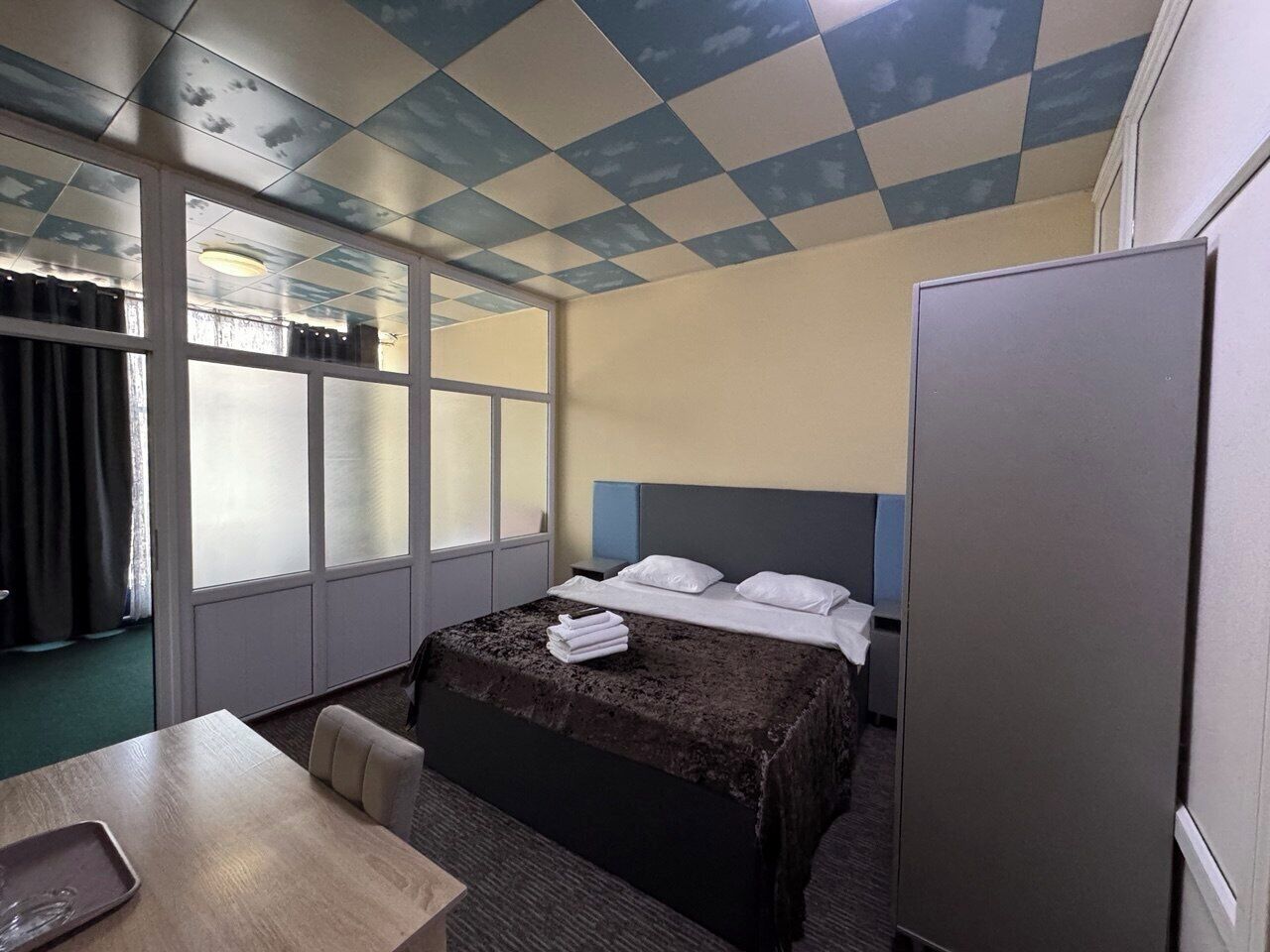 A hotel room with a checkered ceiling, a bed with white linens, a desk with a chair, and a refrigerator.