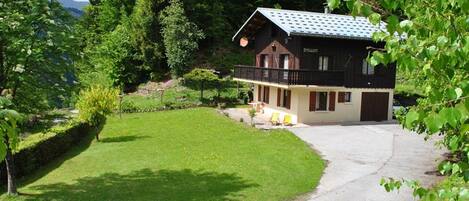 A two-story chalet with a balcony, surrounded by a well-maintained lawn and trees.