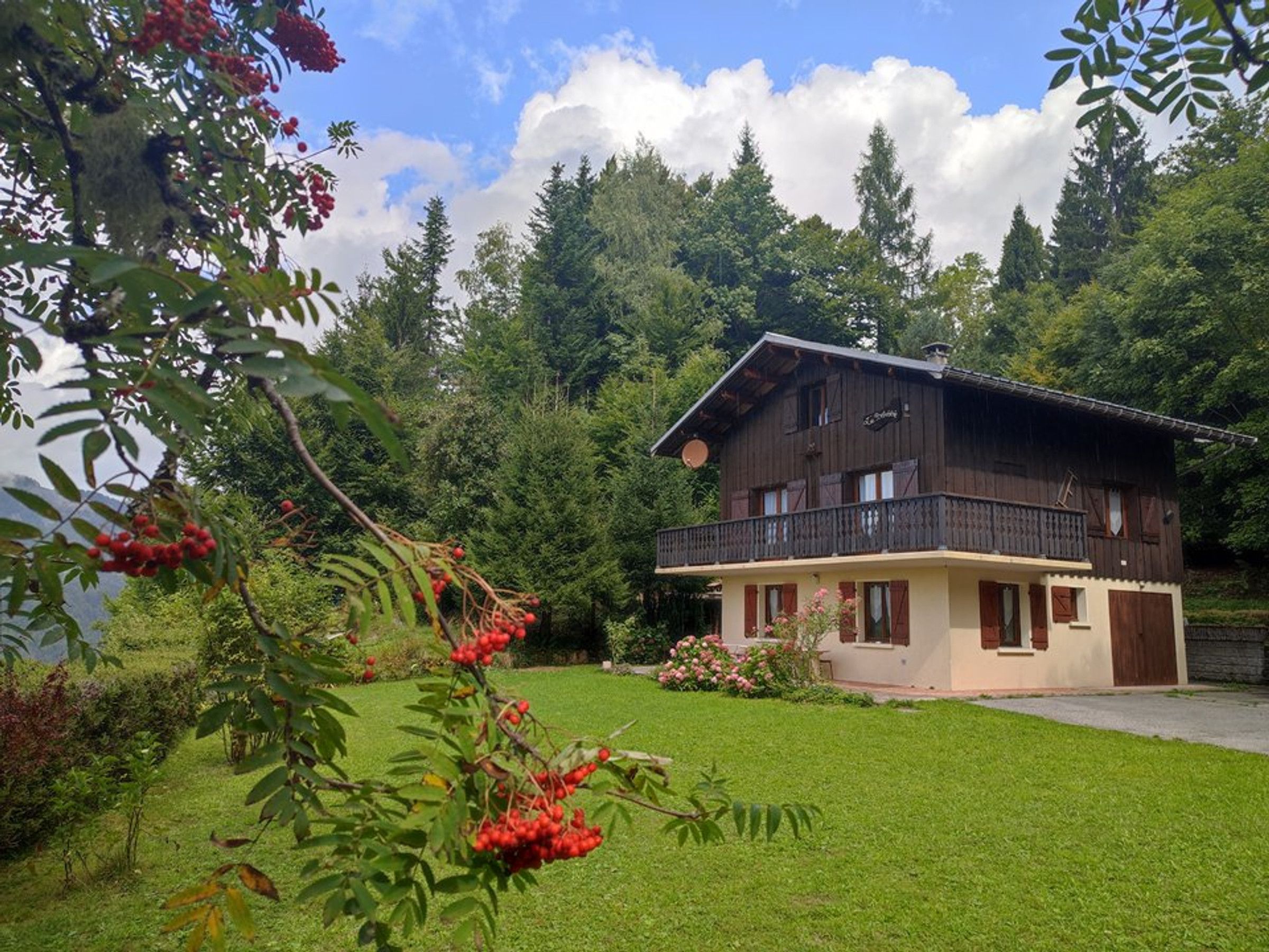 A chalet-style house with a balcony, surrounded by a well-maintained lawn and dense forest.