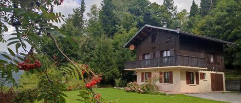 A chalet-style house with a balcony, surrounded by a well-maintained lawn and dense forest.