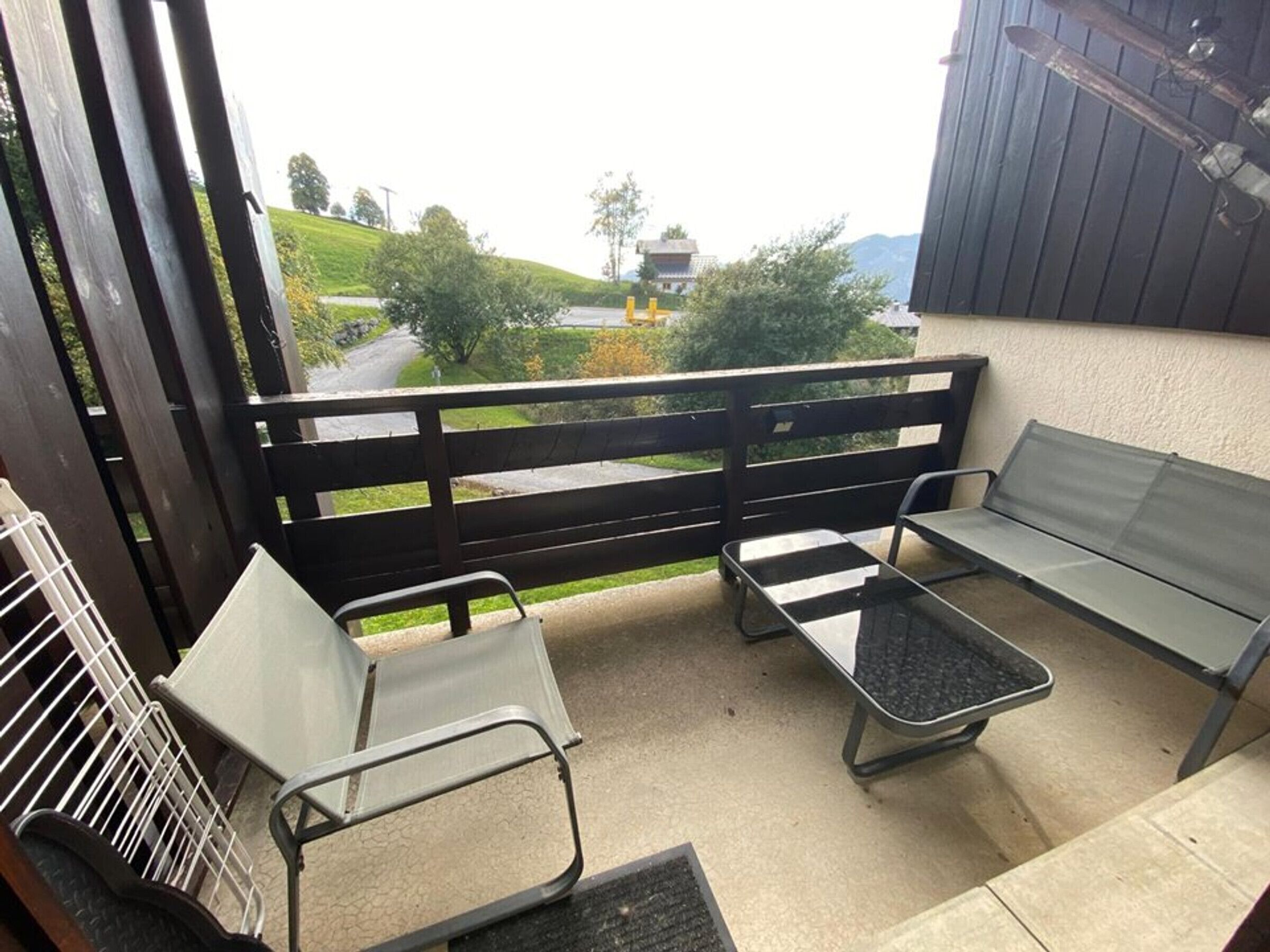 En balkon med gelænder i sort metal, to metalstole, et lille bord og udsigt over et grønt landskab med en vej og bygninger i det fjerne.