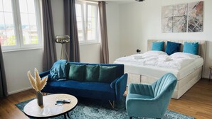 A modern hotel room with a large bed, a blue sofa, a round table with papers, a floor lamp, and a view of the city through the windows. - Schängel Chic apartment centrally located in the old town (Koblenz)