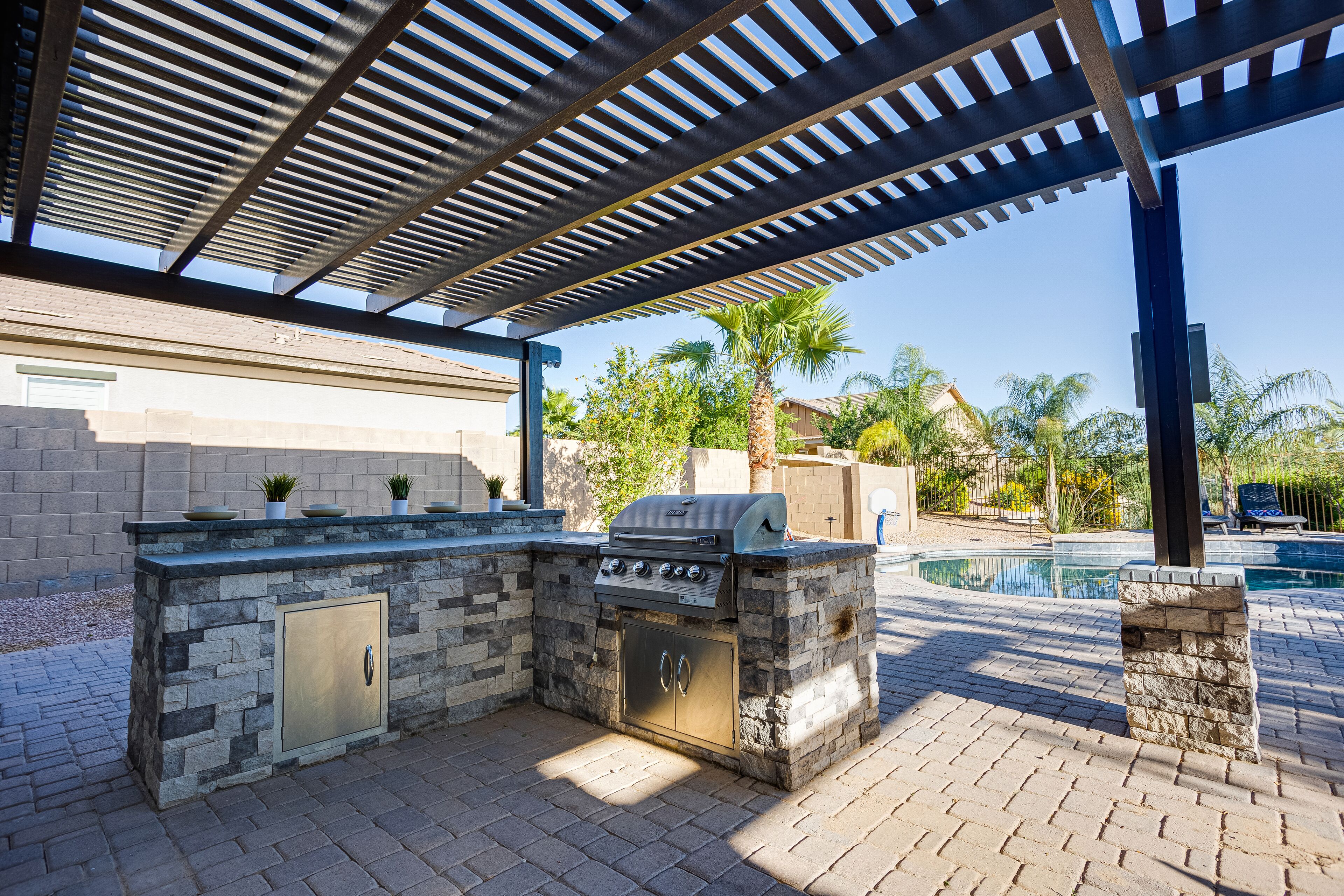 Eine Außenküche mit Grill und Stauraum, unter einer Pergola, in der Nähe eines Schwimmbeckens.