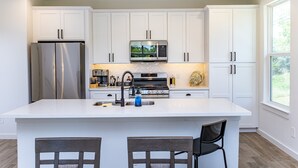Une cuisine moderne avec des meubles blancs, des appareils en acier inoxydable, un îlot central doté de sièges de bar et une vue par la fenêtre.
