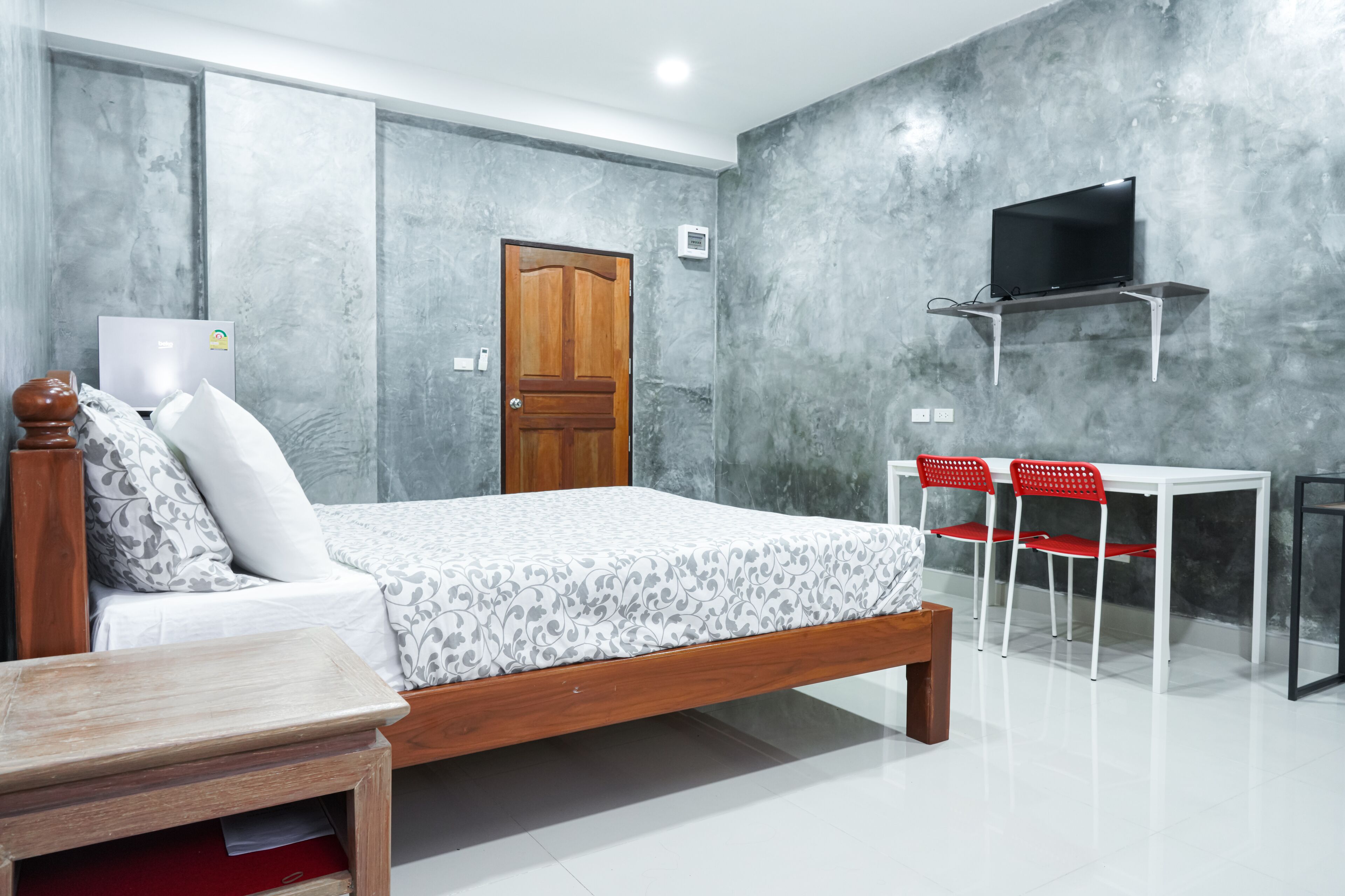 A bedroom with a bed, a wooden nightstand, a desk with two red chairs, a television on a stand, and a door.