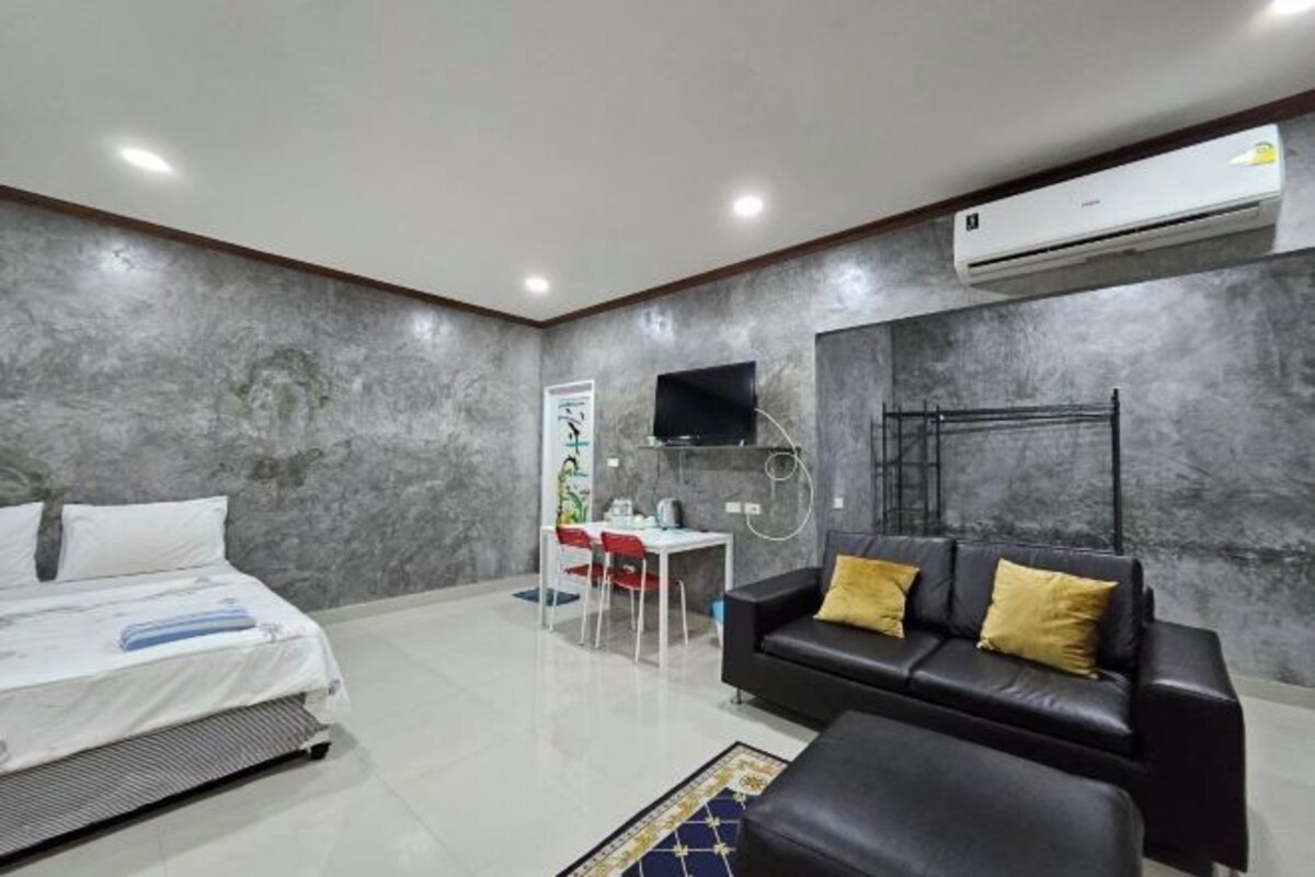 Una habitación de hotel moderna con una cama, un sofá de cuero negro con cojines amarillos, una mesita con una silla y un equipo de aire acondicionado en la pared.
