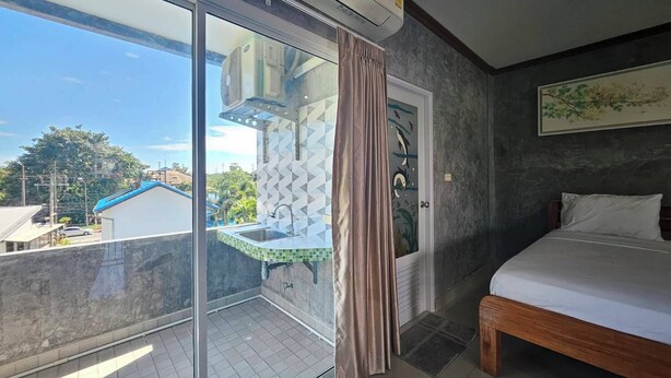 A bedroom with a bed, a window with a view of buildings and trees, a sliding door with a sink, and an air conditioning unit. - Guesthouse Phuket Airport (Sa Khu)
