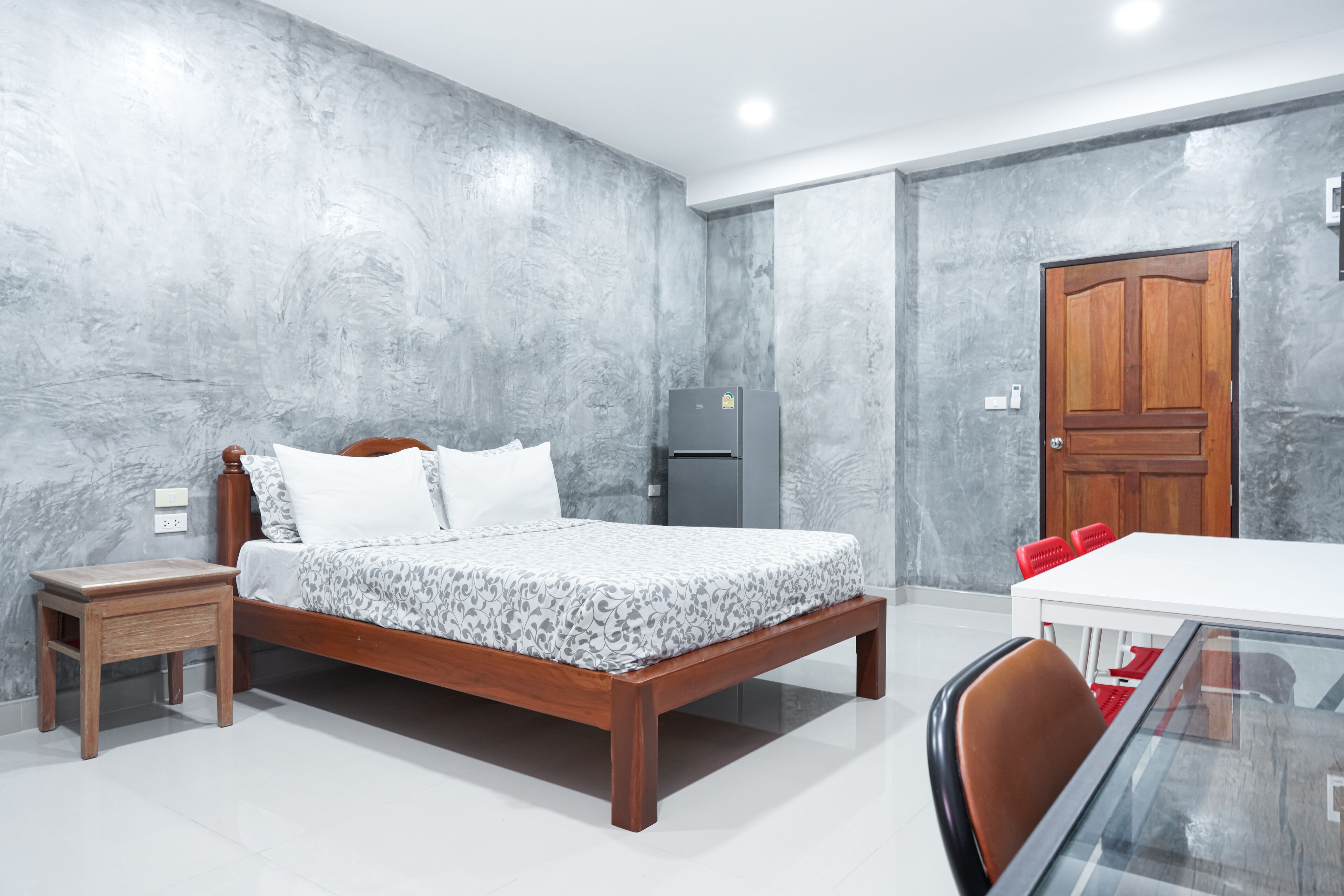 Een slaapkamer met een houten bed, een klein nachtkastje, een koelkast, een houten deur en een eettafel met stoelen.
